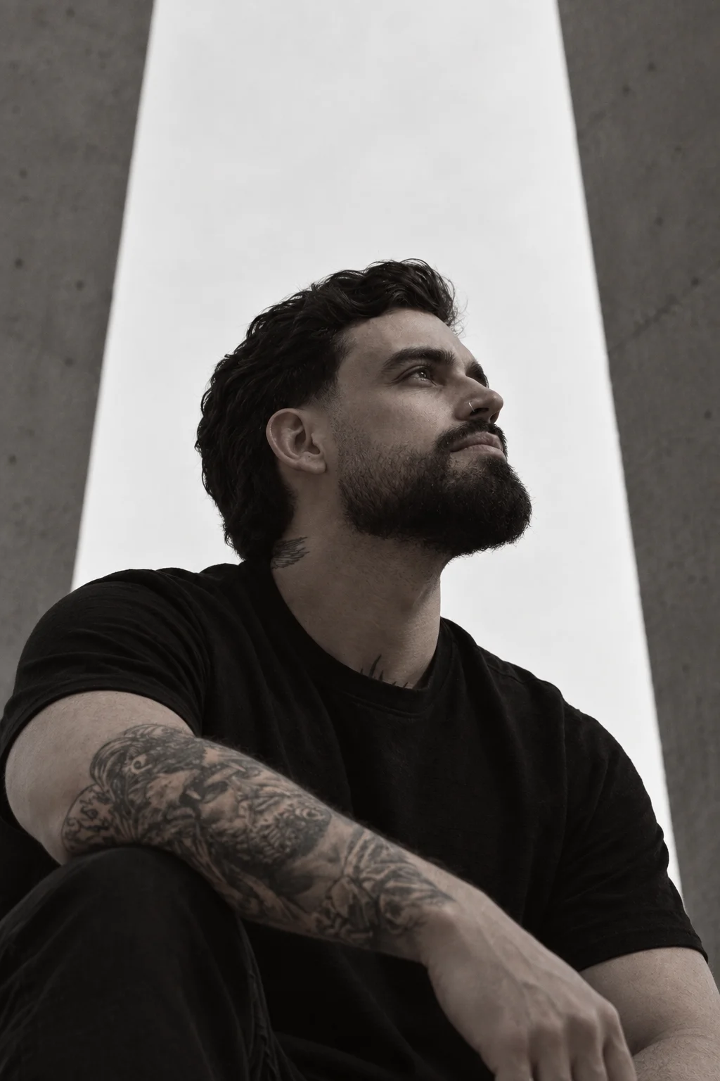 Retrato em preto e branco de Allyson Melo sentado entre dois pilares de concreto, olhando para o alto.